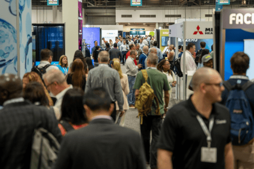 Crowd of business professionals walking through a busy trade show with exhibit booths and signage visible in the background.